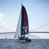 The Plastiki sailing vessel on the water with sails unfurled, in front of a large suspension bridge.