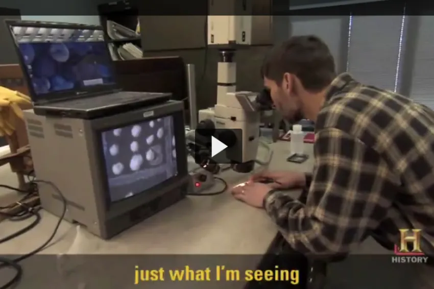 A screenshot from video showing Brian Huber behind a microscope. 