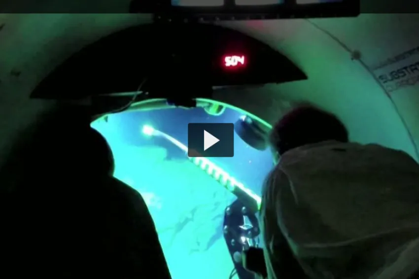 Screenshot from "Deep Reef Observation Project Video" showing two researchers looking out a window of a submersible.