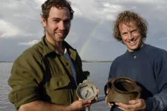 Two men each hold a ceramic vessel at the archaeological excavation