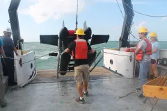 A large, black instrument is hauled onto a ship's deck by researchers wearing hard hats and life vests. 