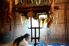 A man, surrounded by fish skins hanging to dry, kneels before a large pit as he demonstrates the tanning process.