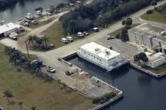 an aerial shot of a marine station with several buildings, cars, palm tree, and a square boat.