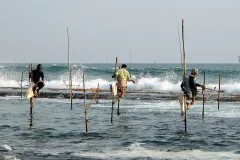 Three fishermen sit on wooden stilts that are sticking out of the ocean as they fish.