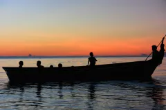 A silhouette of a small boat carrying 8 passengers on tranquil water, with a pink horizon. 