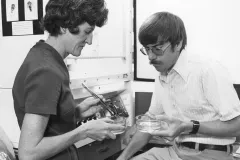 A woman standing and a man sitting on a desk, both looking at specimens