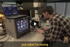 A screenshot from video showing Brian Huber behind a microscope. 