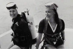 two women sit in scuba gear on the edge of a boat