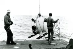 two men on the back deck of a ship pull in a tethered pod