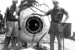 two men stand next to a metal orb on the back deck of a ship