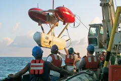 a large red and white autonomous vehicle hangs suspended over the ocean