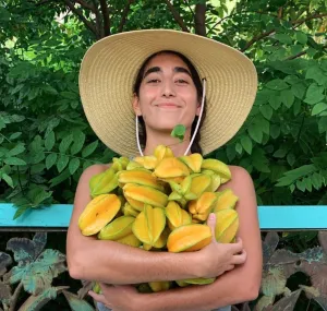 An image of Alia Payne wearing a large straw hat and holding an armful of fruit.