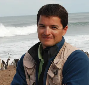 Pablo Garcia Borboroglu standing on the beach with penguins in the background.