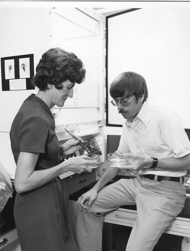 A woman standing and a man sitting on a desk, both looking at specimens