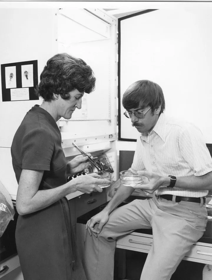 A woman standing and a man sitting on a desk, both looking at specimens