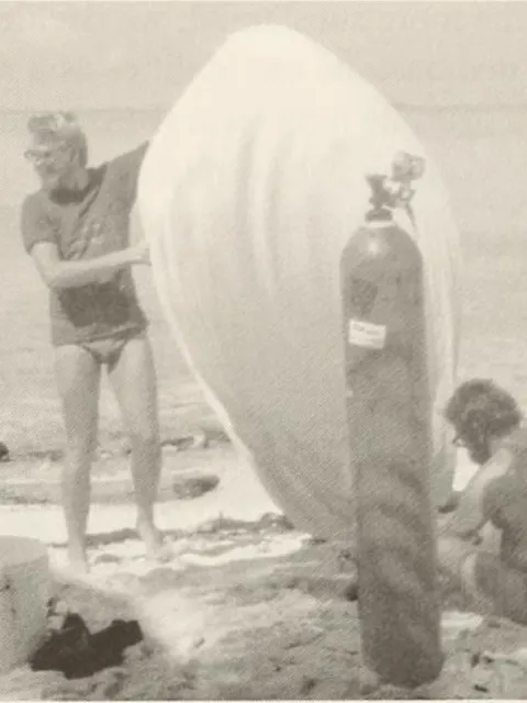 two men work on a beach with a massive, white balloon and helium tank