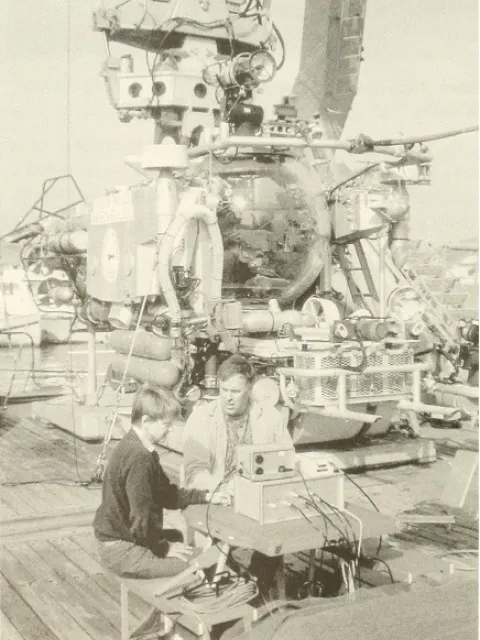 two scientists work on a piece of technology on a ship deck with a submersible in the background